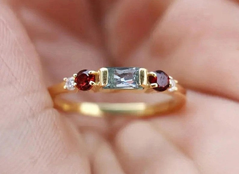 A gold ring with a rectangular clear center stone and two round red side stones, shown resting on a person's fingers.