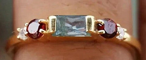 A close-up of a gold ring worn on a finger, featuring a rectangular light blue gemstone in the center, flanked by two round dark red gemstones and small clear accent stones.