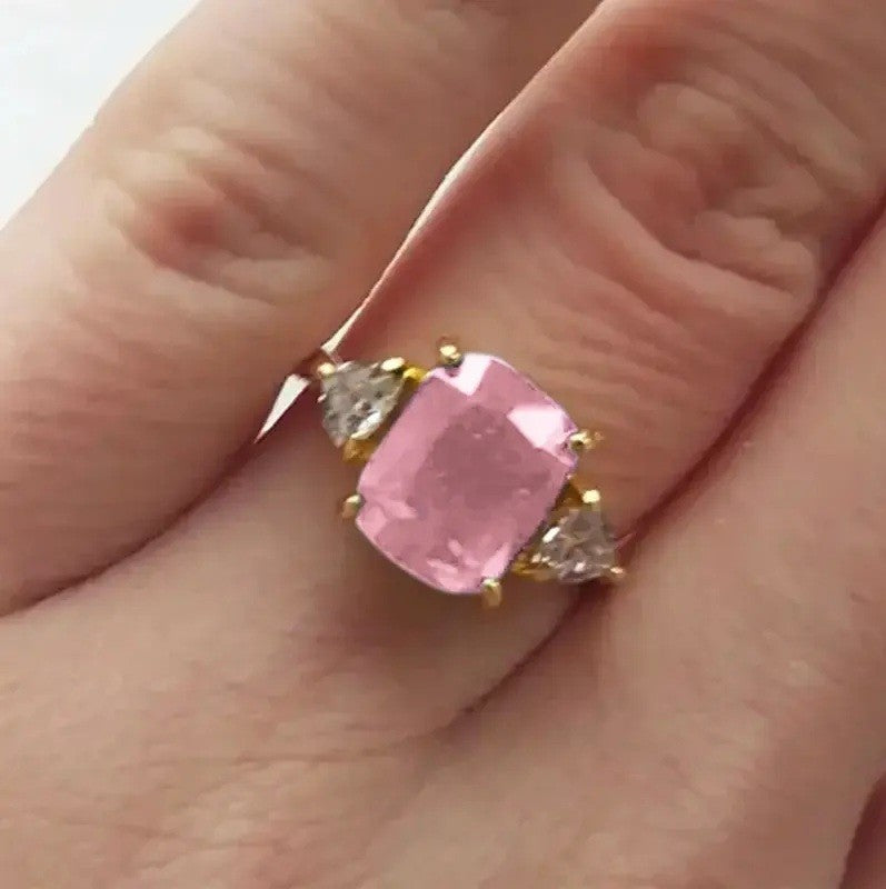 A close-up of a gold ring with a large rectangular pink gemstone and two small clear accent stones, worn on a person's finger.