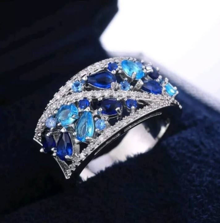 A wide silver-toned ring featuring marquise and oval-cut blue sapphires and clear stones, displayed on a dark velvet jewelry box.
