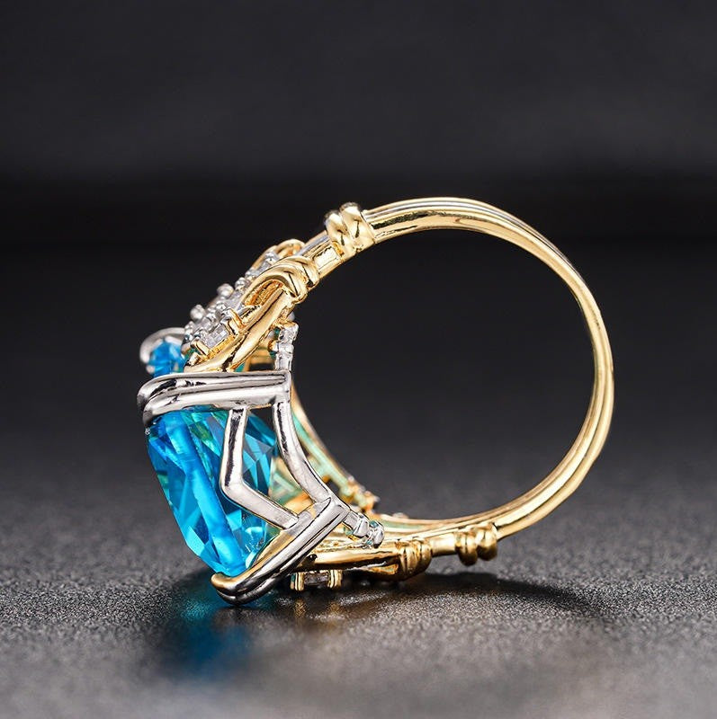 A gold and silver ring featuring a large, vibrant blue marquise-cut gemstone, accented with smaller clear stones, shown on a dark textured background.