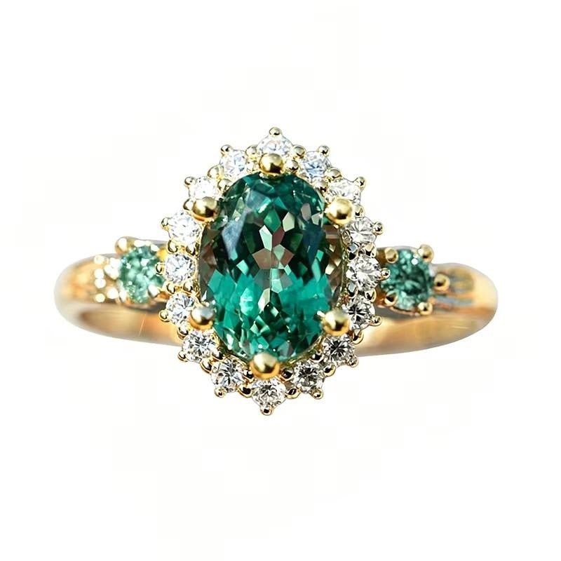 A yellow gold ring featuring a large oval-cut green emerald gemstone in a halo setting, accented by smaller round green and clear stones on a plain white background.