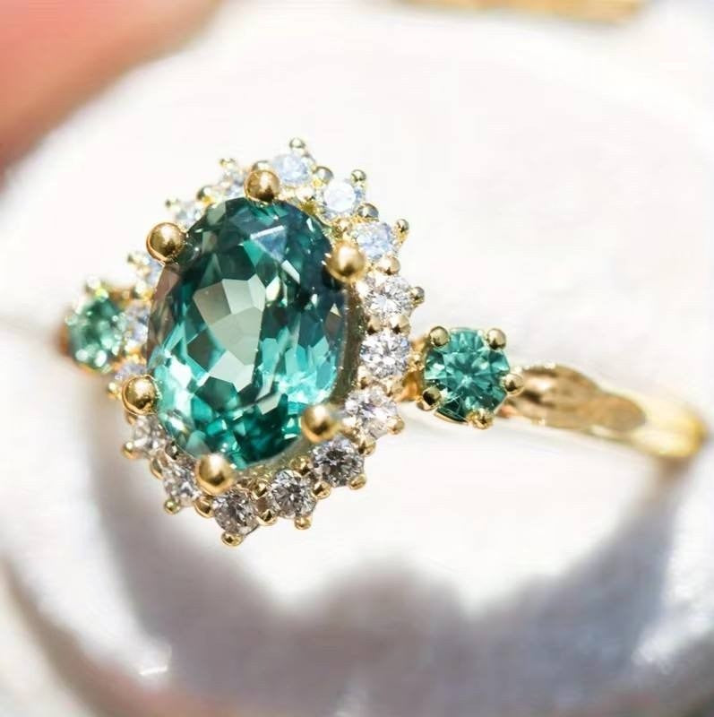 A yellow gold ring featuring a large oval-cut green emerald gemstone, surrounded by a halo of small clear stones, with two smaller green gemstones on the band, shown resting on a white surface.