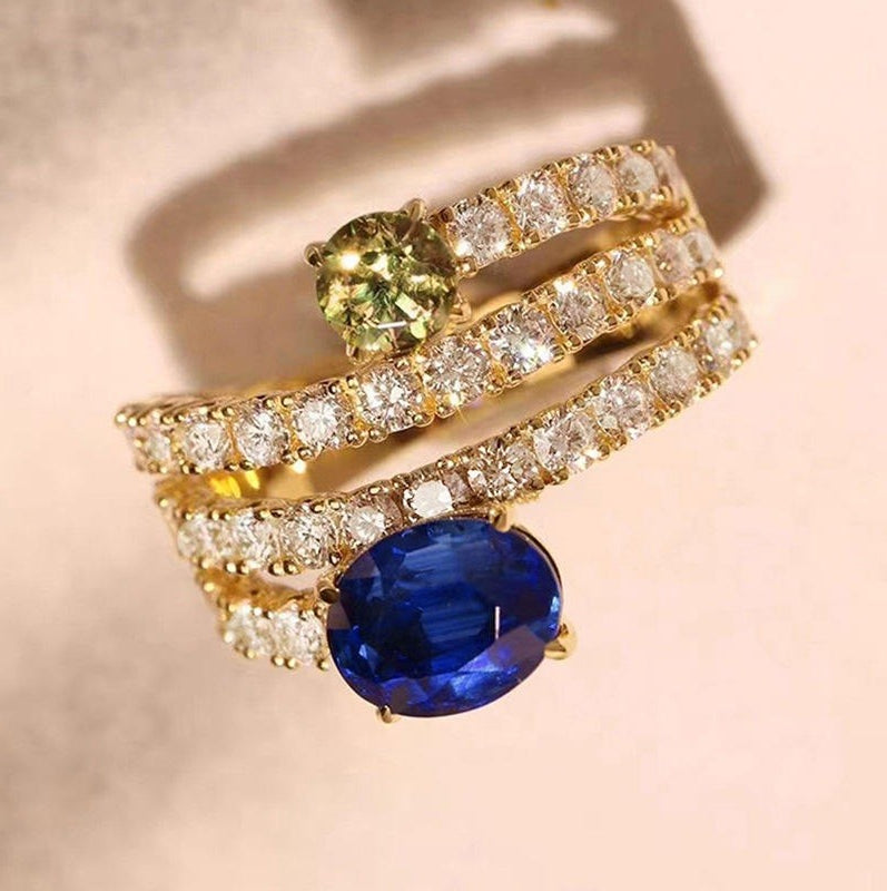 A gold ring set with a large oval blue sapphire and a smaller oval green peridot, accented with multiple rows of round diamonds, photographed on a soft beige background.