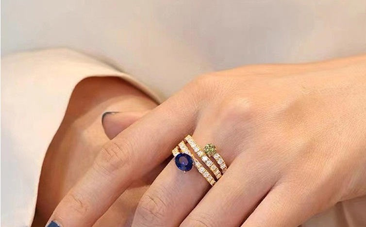 A close-up of a hand wearing a stack of gold rings, including a ring with a round blue gemstone and two bands set with small clear stones, against a soft cream-colored background.