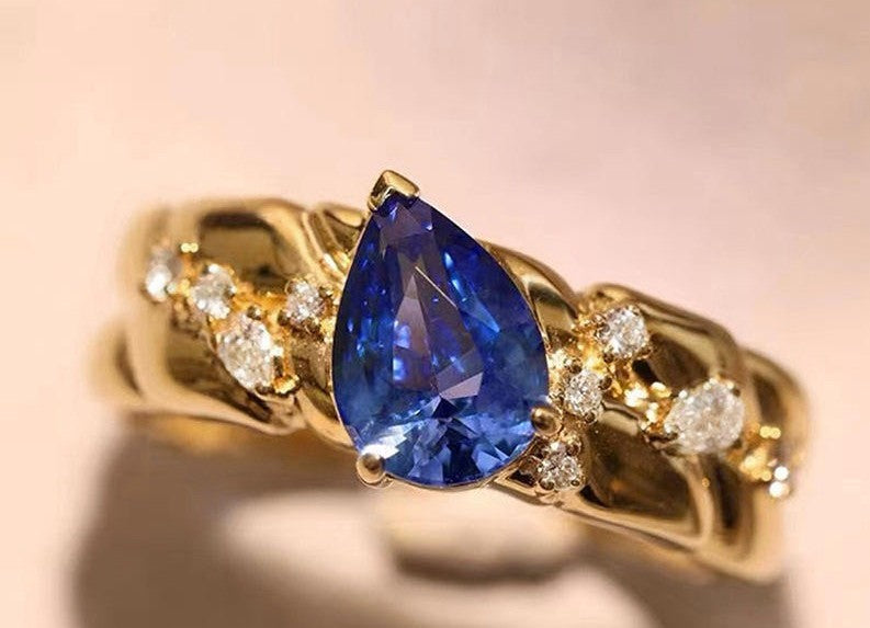 A gold ring featuring a large pear-cut blue sapphire center stone, accented with small round diamonds on a soft beige background.