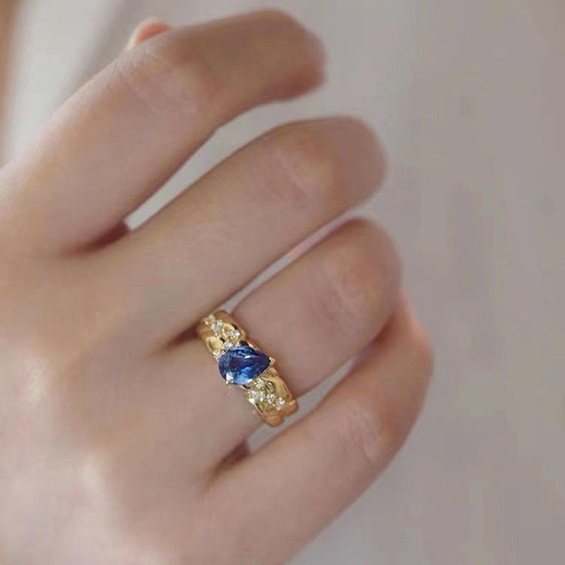 A gold ring with a central blue gemstone and small clear accent stones, worn on a person's finger against a soft light background.