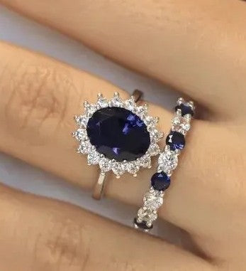 A close-up of a hand wearing two rings: a larger silver ring with an oval deep blue gemstone surrounded by small clear stones, and a matching slim silver band with repeating blue and clear stones.