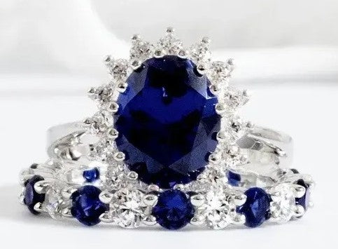A silver-toned ring featuring a large oval deep blue gemstone surrounded by a halo of small clear stones, with a matching band decorated with smaller oval blue gemstones and clear accents, set against a soft white background.