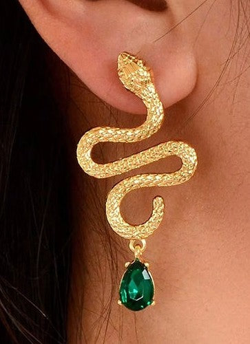 A close-up of a gold snake-shaped dangle earring worn on a person's ear, featuring a textured gold snake body and a dangling teardrop-shaped green gemstone.